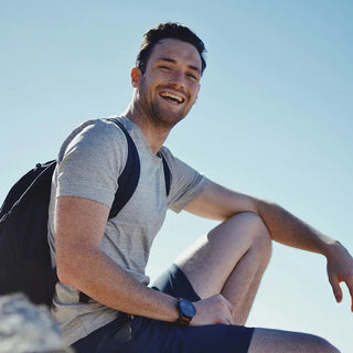 Un hombre con camiseta gris, pantalones cortos y mochila está sentado al aire libre sobre una roca y sonríe a la cámara bajo un cielo azul claro.