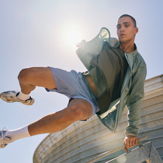 Eine Person in sportlicher Kleidung springt über ein Geländer im Freien, mit einem runden modernen Gebäude und hellem Sonnenlicht im Hintergrund.