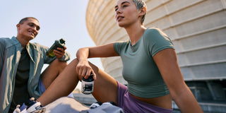 Dos personas con ropa deportiva se relajan al aire libre cerca de un edificio moderno, sosteniendo botellas de suplementos y bebidas después de un entrenamiento.