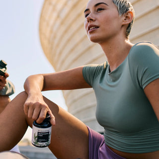 Una persona con ropa deportiva sentada al aire libre sosteniendo un envase etiquetado "Performance Capsules", con un edificio moderno y curvado de fondo.