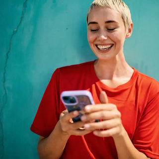 Eine Person mit kurzen blonden Haaren und einem roten Hemd steht lächelnd vor einer blaugrünen Wand und benutzt dabei ein Smartphone.
