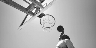Una persona a punto de encestar una canasta al aire libre contra un cielo despejado vista desde abajo.
