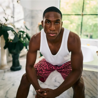 Un hombre con una camiseta blanca sin mangas y shorts rosas con estampado de leopardo está sentado con las manos cruzadas sobre un suelo de mármol en el interior. En el fondo, hay grandes ventanas y plantas.