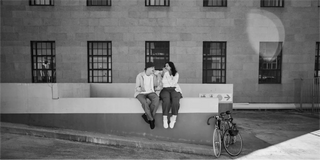 Dos personas sentadas en un muro bajo junto a una bicicleta frente a un edificio con varias ventanas.