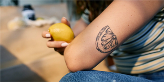 Persona sosteniendo un limón con un tatuaje de rodaja de limón en su antebrazo; usando una camisa de rayas y jeans.