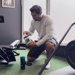 Un hombre se arrodilla en el suelo de un gimnasio, vertiendo polvo en una botella mezcladora junto a una máquina de remo.