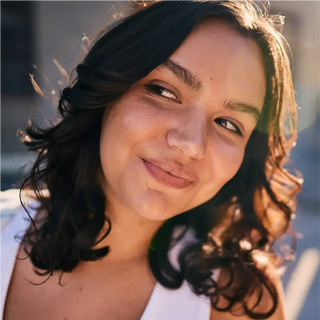 Una persona con cabello oscuro y ondulado y una camiseta blanca sonríe ligeramente mientras mira de lado al sol.