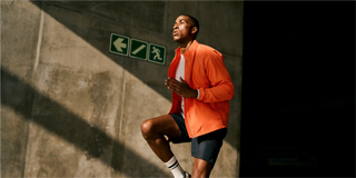 Un hombre con ropa deportiva que está corriendo dentro de un edificio junto a una pared de concreto con carteles de salida y escalera iluminados por la luz del sol.