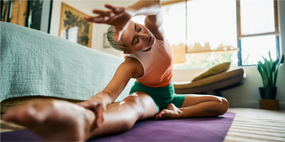 Persona sentada en una esterilla de yoga en una sala de estar, estirando un brazo sobre su cabeza hacia su pierna extendida.