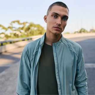 Un joven con el pelo corto que lleva una chaqueta azul claro sobre una camiseta oscura está de pie al aire libre en una zona pavimentada con árboles desenfocados en el fondo.