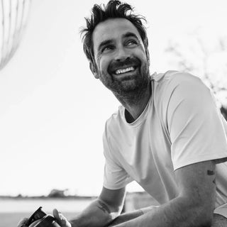 Un hombre con barba y cabello despeinado está sentado al aire libre, sonriendo y sosteniendo una cámara. Lleva una camiseta de color claro. La imagen es en blanco y negro.
