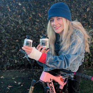Una mujer rubia equilibra dos botellas sobre su bicicleta, rodeada de elementos naturales.